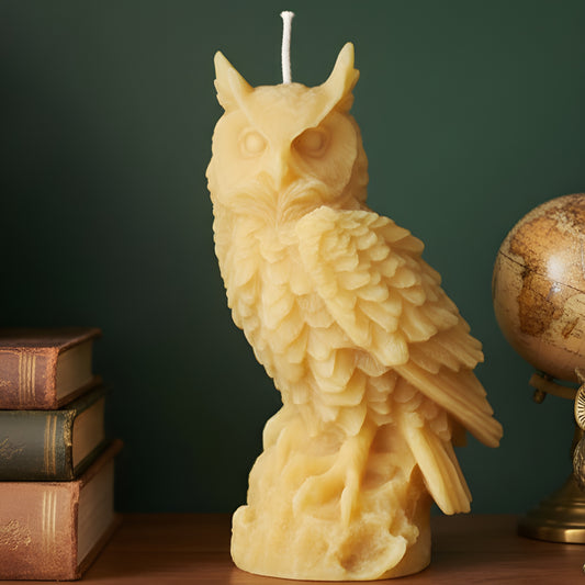Tall golden owl beeswax candle with layered feathers beside books against a green wall.