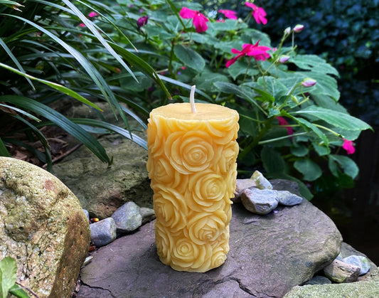 Yellow beeswax pillar covered in small rose shapes, displayed on a stone in a flower garden.