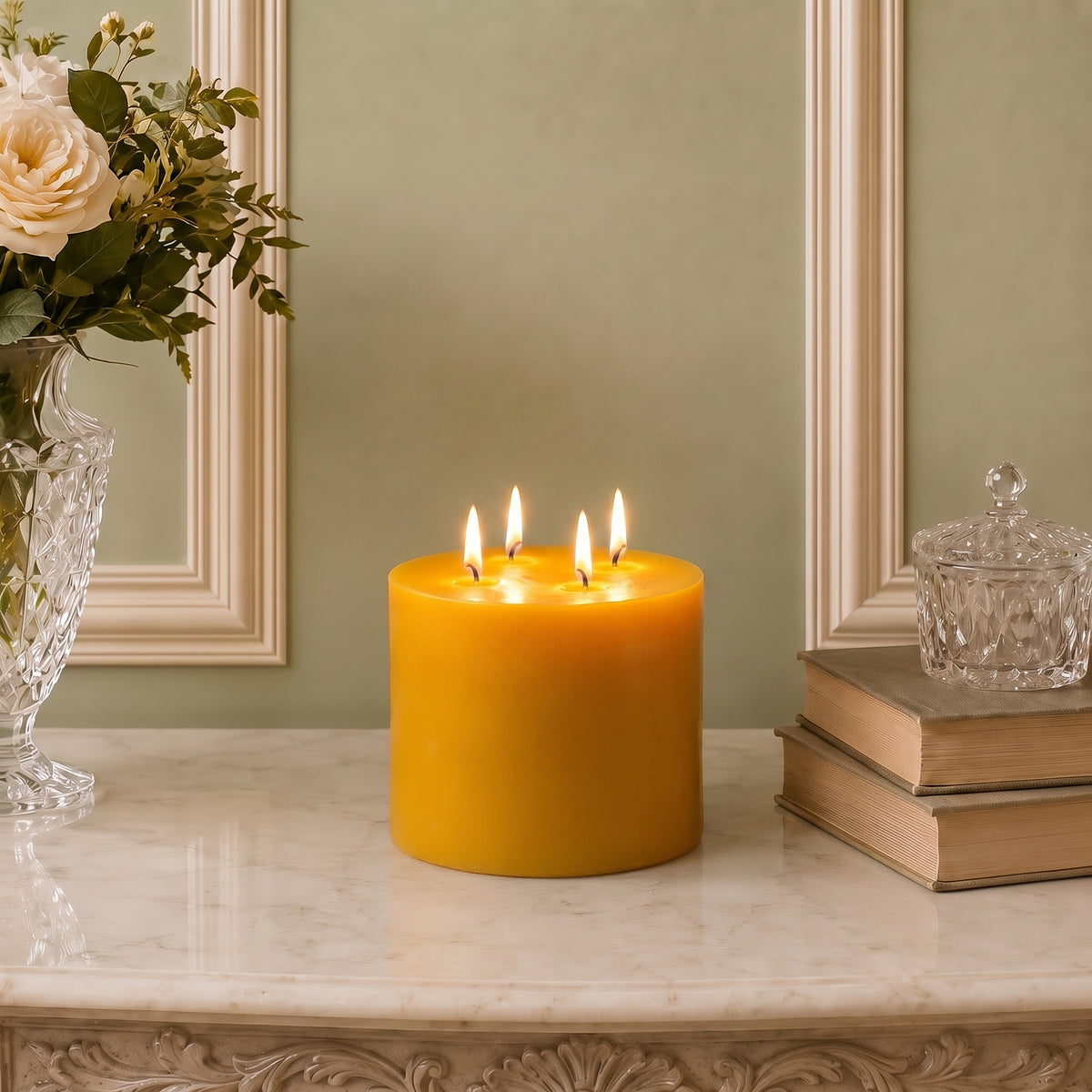 Golden four-wick beeswax pillar on a pale mantel beside flowers and books.